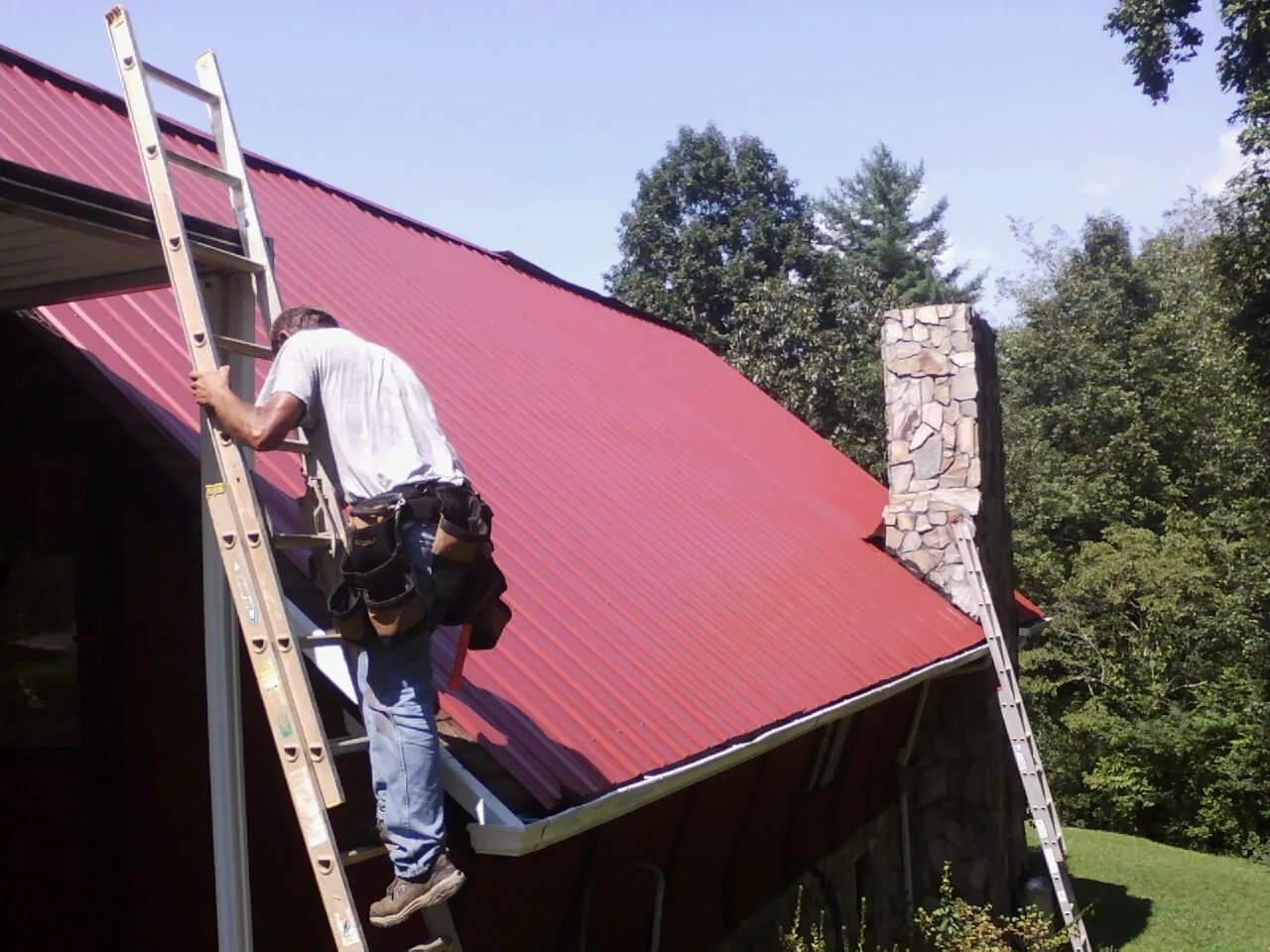 Roofer on a ladder beside a red metal roof and stone chimney — Soffit & Fascia Repair by Roofing Brothers in Gilmanton Iron Works